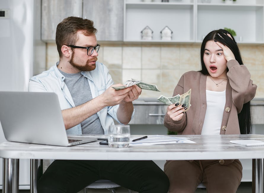 A man with glasses and a beard is seated at a table inside a home, handing over dollar bills to a woman with long black hair, who appears surprised and distressed, with her hand on her forehead. The table has a laptop, a glass of water, a pen, and some papers. In the background, there is a modern kitchen with light-colored tiles and white shelves decorated with small houseplants and picture frames. The scene captures a moment of financial disagreement related to house removals or moving costs, with the context suggesting concerns over hidden fees or unexpected expenses during a home relocation. The environment is well-lit, and the visible objects include cardboard boxes and packing materials on the table, hinting at ongoing packing and moving preparations overseen by Merton Movers, a removals service provider.