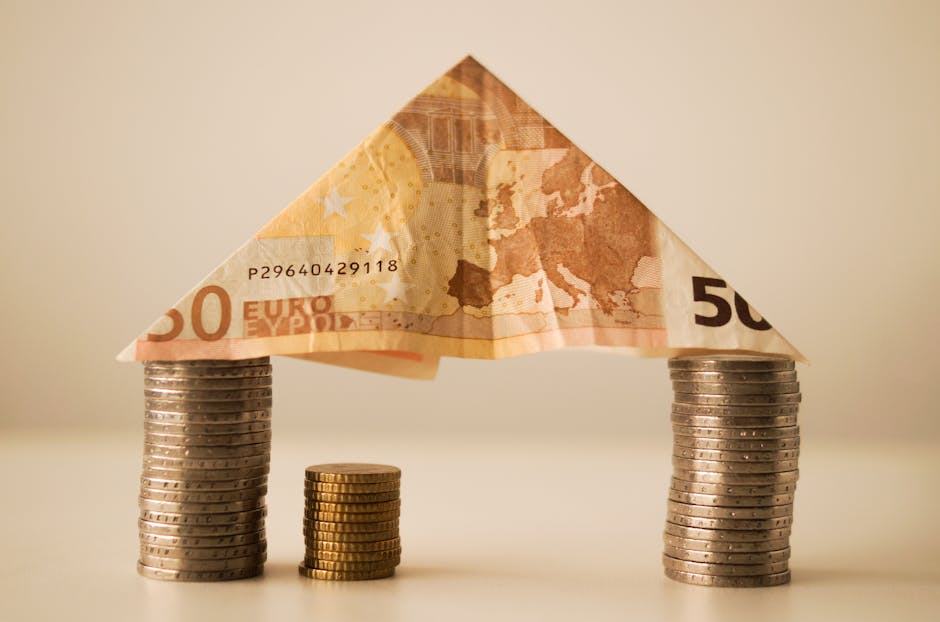 A creative arrangement depicts a house structure formed by a folded 50 euro banknote placed atop two stacks of silver coins representing support pillars. A smaller stack of gold coins stands in the middle, serving as a base for the banknote. The scene is set on a light surface with a neutral background, highlighting the materials used in packing and transporting household items. The coins and banknote suggest concepts related to home relocation, furniture transport, and moving logistics, aligning with professional removals services offered by Merton Movers. The lighting is soft and even, emphasizing the textures of the coins and the detailed design of the banknote, illustrating the complex process of packing, loading, and transporting belongings during a house removal.