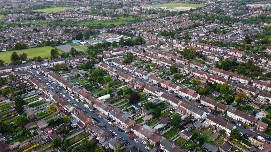 An aerial view of a residential neighbourhood showing rows of terraced houses with front gardens and driveways, parked cars lining the streets, and trees interspersed among the properties. The scene captures an ongoing home relocation process, with Merton Movers' van visible near the pavement, where packing materials such as cardboard boxes and furniture blankets are arranged for loading. In the background, open green spaces and sports fields are visible, indicating a suburban area with a mix of residential and recreational land. The image highlights the organized environment typical of estate-by-estate moves in Colliers Wood to Mitcham, illustrating the logistics and careful handling involved in household furniture transport and packing during house removals.