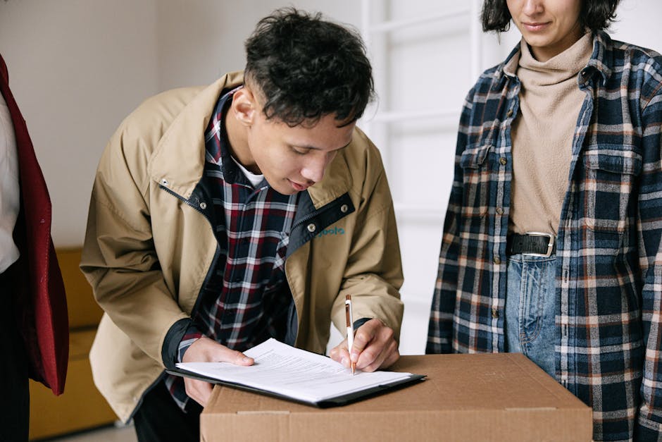 A man wearing a beige jacket over a red, blue, and white checkered shirt is leaning over a cardboard box placed on a table or countertop. He is holding a black pen and appears to be filling out or signing a document on a clipboard. The woman standing next to him is dressed in a plaid shirt over a beige turtleneck and high-waisted blue jeans, with her hands by her sides. The background shows a white wall, possibly in a home or office setting, with some shelving or storage units partially visible. The scene depicts a packing and moving process, with attention to proper documentation, typical of home relocation or furniture transport activities, which are services offered by Merton Movers. The lighting is bright and even, emphasizing the focus on the individuals and the packing materials involved in the move.