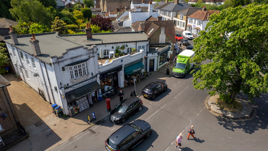 An aerial view of a residential neighbourhood showing rows of terraced houses with front gardens and driveways, parked cars lining the streets, and trees interspersed among the properties. The scene captures an ongoing home relocation process, with Merton Movers' van visible near the pavement, where packing materials such as cardboard boxes and furniture blankets are arranged for loading. In the background, open green spaces and sports fields are visible, indicating a suburban area with a mix of residential and recreational land. The image highlights the organized environment typical of estate-by-estate moves in Colliers Wood to Mitcham, illustrating the logistics and careful handling involved in household furniture transport and packing during house removals.