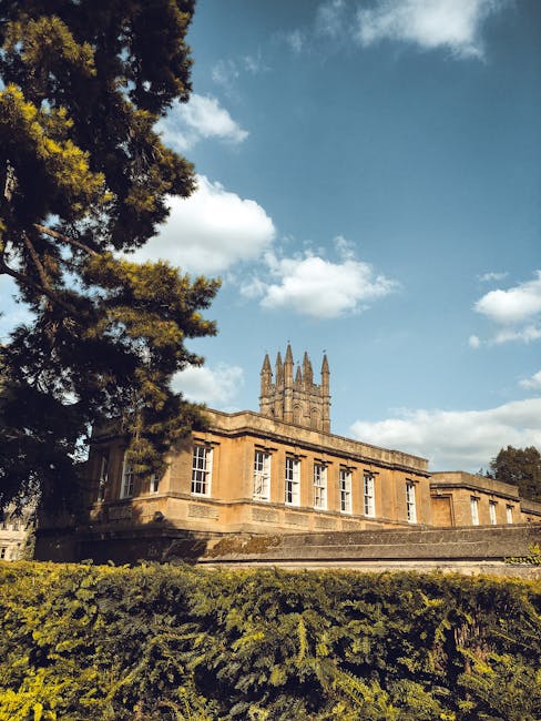 A historic sandstone building with large windows and ornate architectural details, situated on a raised platform with steps leading up to the entrance. To the left, a tall evergreen tree with dense foliage partially obscures the view, casting shadows on the building's facade. The sky above is partly cloudy with patches of blue, and the scene is illuminated by natural daylight. In the foreground, a well-maintained hedge runs along the base of the building, with the surrounding area appearing calm and tidy. This setting suggests a location in a park or institutional grounds, relevant to discussions about property, estate management, or relocation logistics associated with house removals and moving services, with the presence of an historic building adding context to the environment often involved in property transactions or venue considerations.