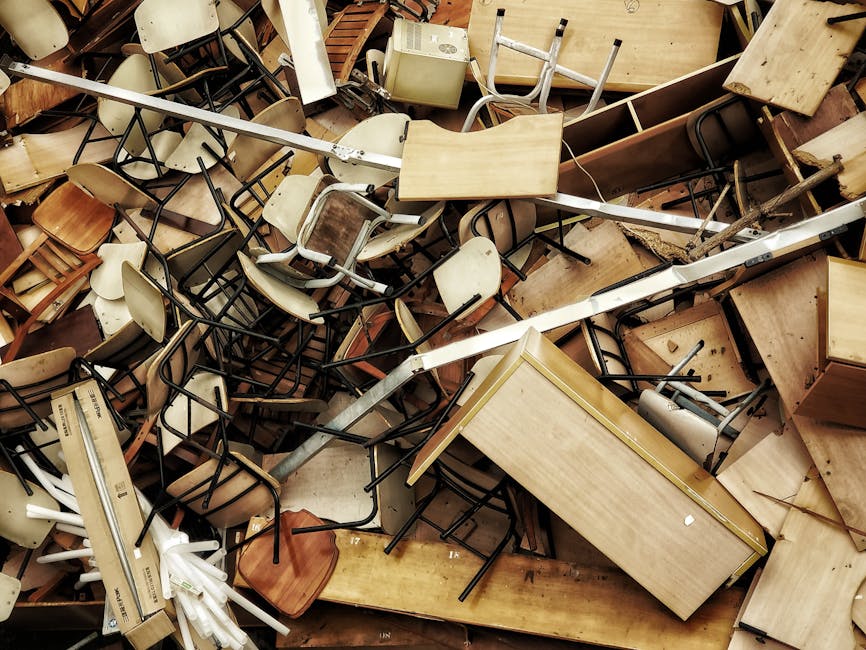 A large pile of discarded furniture and household items, including wooden chairs, tables, and cabinets, mixed with plastic and cardboard packaging materials. The items are randomly stacked inside a property, with some furniture partially broken or damaged, and a metal trolley or hand truck visible among the clutter. The background shows a doorframe and a concrete or tiled floor, suggesting an indoor setting in the process of clearance or removal. The scene captures a cluttered environment typical of furniture disposal during home relocation or professional removals, with materials prepared for disposal or recycling. Merton Movers’ removal service is relevant to this context, supporting legal disposal options for unwanted furniture to avoid fines.