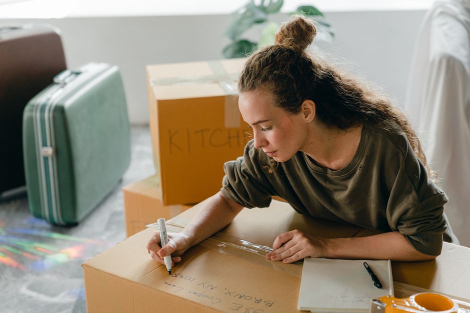 A woman with curly brown hair tied up in a bun is leaning over a large cardboard box inside a room, writing with a black marker on the top of the box. She is wearing a brown sweatshirt and appears focused on labeling the box for a house removal process. Behind her, there are additional packing materials, including a green hard-shell suitcase with a handle and wheels, and a cardboard box with the word 'KITCHEN' written in red marker. The room has natural lighting coming from a window, with a light-colored wall and some green plants visible in the background. The scene captures part of the loading or packing stage of a home relocation where boxes and luggage are prepared for transportation. Merton Movers, a professional removals company, provides services related to these moving logistics, including packing and furniture transport, often requiring permits during the moving day process in Merton, as referenced in the webpage titled 'Do I need a permit from Merton Council for moving day?'