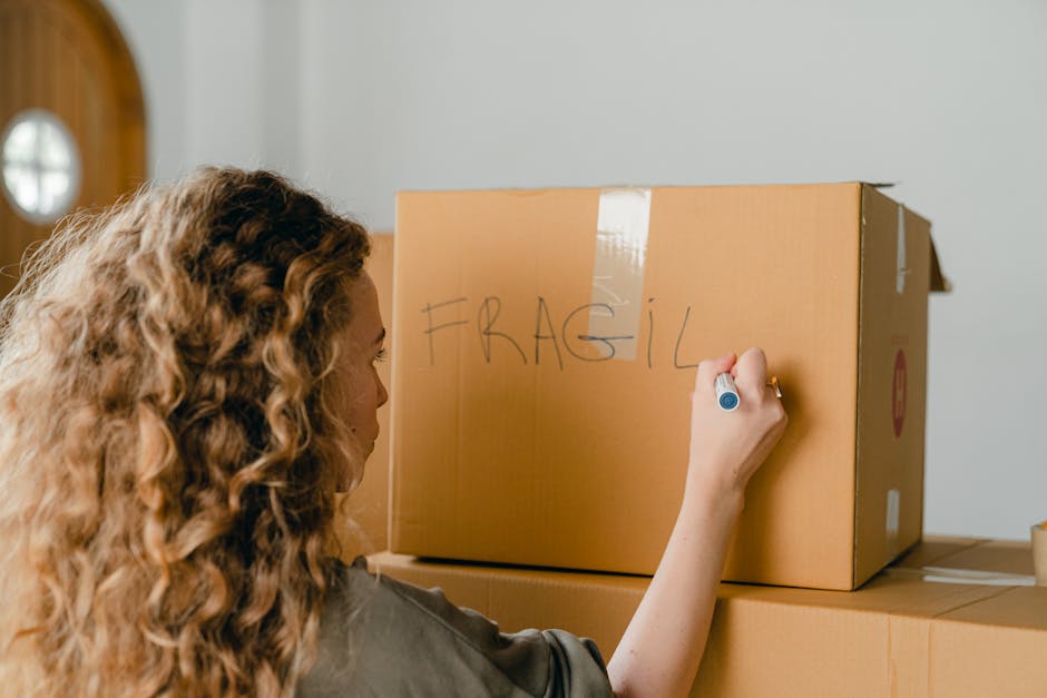 A woman with curly, light brown hair is indoors, writing the word 'FRAGIL' in capital letters on a large cardboard box with a blue marker. The box is positioned on a stack of other cardboard boxes, which are part of a home relocation or packing process. The box she is labeling appears sturdy, with brown paper and white tape sealing its top edges. Additional boxes and packing materials are visible in the background, indicating a busy packing environment associated with moving or furniture transport. This scene captures the careful handling and organization involved in packing fragile items during a house removal, reflecting professional moving practices supported by Merton Movers, a specialist removals company. The lighting is natural, highlighting the preparation steps involved in household relocation and ensuring items are properly labeled for transport and handling.