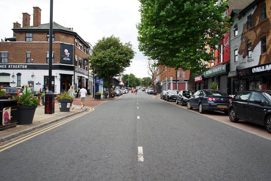 A street scene on Wimbledon High Street featuring a row of parked cars on the right side of the road, including compact and hatchback models, with a few pedestrians walking along the pavement. On the left, outdoor seating with large black planters filled with colorful flowers is visible outside a café or restaurant, with a few customers seated. The buildings have a mix of brick and painted facades, housing various shops and businesses such as a pharmacy and a pub. In the foreground, a partially visible black van belonging to Merton Movers is parked near the curb, adjacent to a loading bay area marked for deliveries and moving logistics. The street is lined with mature trees providing shade, and the overall setting appears calm and typical of a retail and dining district. This scene illustrates an urban environment suitable for home relocation and furniture transport services within the area.