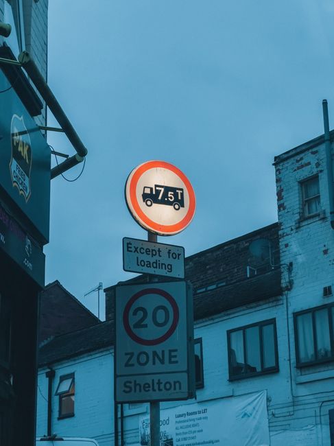 A set of three traffic and parking restriction signs mounted on a metal pole against a backdrop of buildings on Wimbledon High Street during daytime. The top sign indicates a maximum vehicle weight of 7.5 tonnes, with a note below that states 'Except for loading.' Beneath it, a circular sign with a red border displays a speed limit of 20 miles per hour, and below that, a rectangular sign reads 'ZONE.' At the bottom, another sign specifies 'Shelton,' indicating the zone or area name. The buildings in the background include residential or commercial properties with windows, some of which have visible curtains. The sky is overcast, suggesting cloudy weather. This scene depicts typical traffic signage relevant to local vehicle movement regulations, which Merton Movers, specialists in house removals and packing, must consider during the loading and transportation stages of home relocation and furniture transport in the area.
