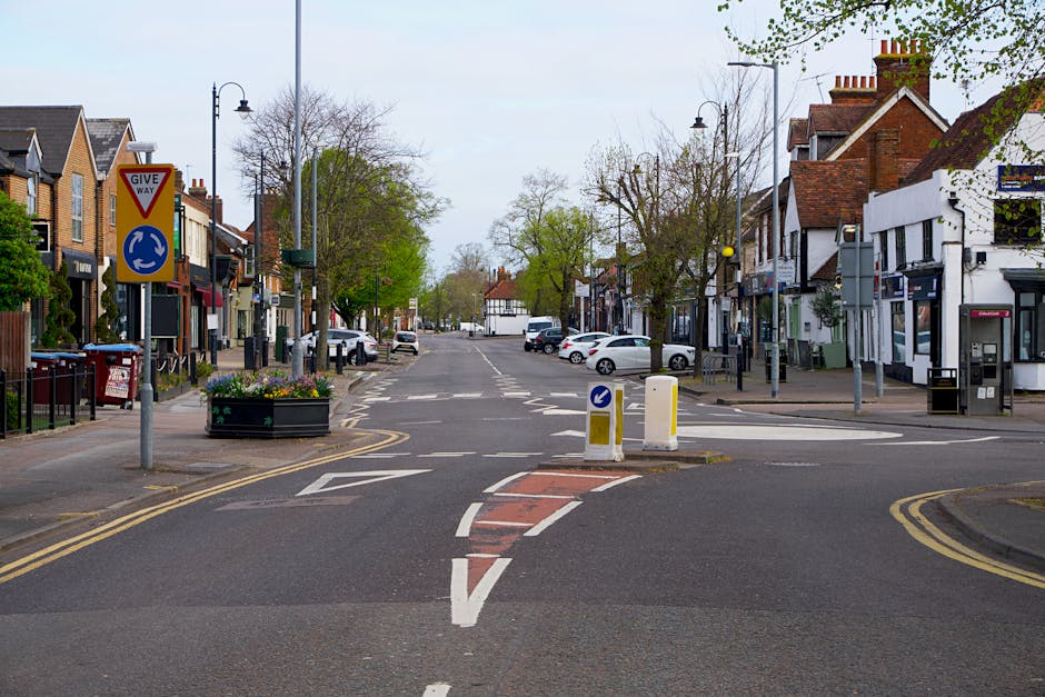 A quiet residential street in Morden with a double mini roundabout, traffic signs including a give way and directional arrow, and a row of houses and shops on either side, some with front gardens. The