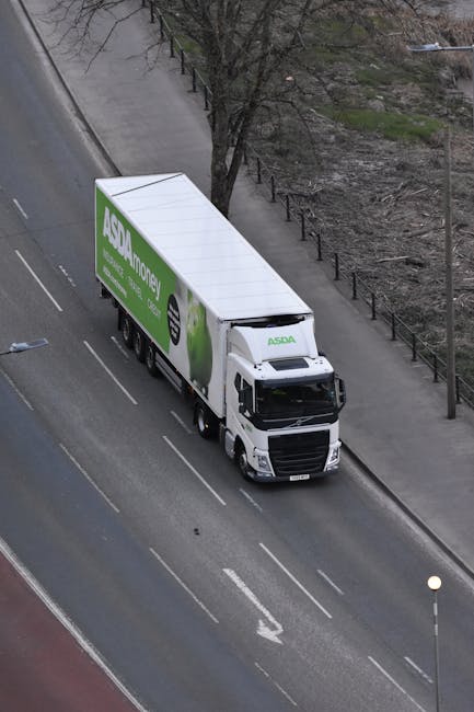 An aerial view of a white ASDA home delivery truck parked on the side of a residential street with narrow driving space in Raynes Park (SW20), Merton. The truck is positioned parallel to the pavement, which is bordered by a row of metal fencing and a tree with bare branches. The truck features a large, rectangular cargo area with the green and white ASDA logo and branding, indicating it is used for furniture transport and home relocation services. The street has marked lanes, with a white dashed line separating the parking area from the driving lane. Nearby, a small streetlamp is visible, and the surrounding environment includes some patches of grass and an area with bare soil or low vegetation, reflecting the urban setting. This scene captures the logistical aspect of moving and transportation within a compact residential area, aligning with professional removals services offered by Merton Movers.