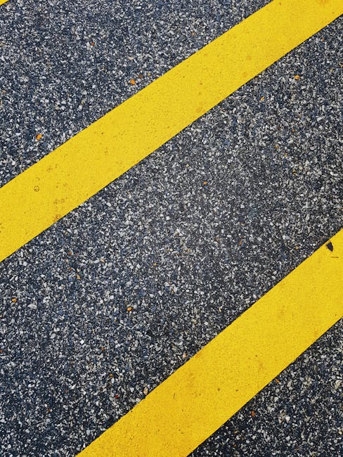 A close-up view of a rough asphalt surface featuring two parallel yellow lines painted across the road, which are commonly used to indicate traffic lanes or caution zones, with small gravel and debris scattered on the surface. In the background, part of a white-painted curb and a small section of pavement leading to a residential property are visible. The yellow lines are slightly worn, and the overall scene suggests a narrow driveway or street where home relocation and furniture transport activities by Merton Movers might take place, especially when navigating tight urban or suburban settings for house removals within Raynes Park (SW20) or similar areas.