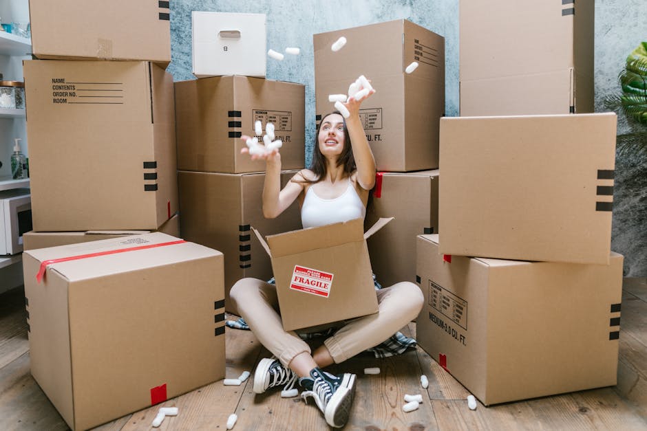 A young woman sitting cross-legged on a wooden floor inside a home surrounded by large cardboard moving boxes and packing materials, with some boxes sealed with red tape and others open, revealing packing foam and items inside. She is wearing casual clothing, including a white tank top, beige pants, and sneakers, and is smiling while throwing packing foam pieces into the air. The background includes a blue textured wall, a small potted plant, and a white kitchen counter with a microwave. This scene illustrates a home relocation process involving packing and furniture transport, as undertaken by Merton Movers, a professional removals company based in Wimbledon, with the focus on efficient packing and loading procedures.