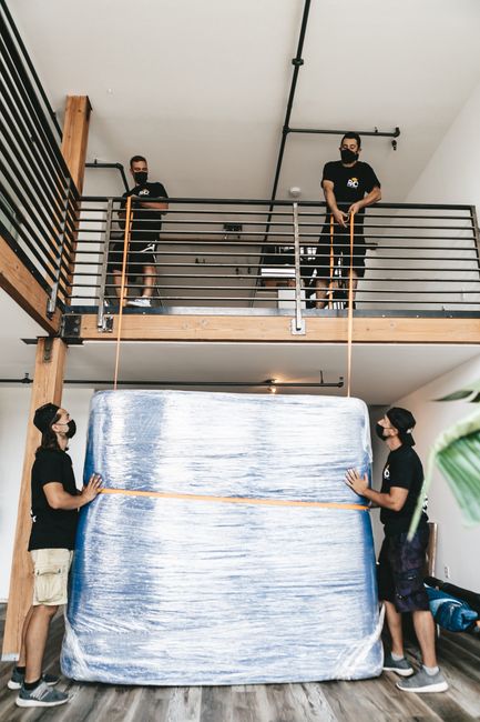 Two movers from Merton Movers, wearing black uniforms and masks, are indoors assisting with a home relocation. They are positioned on the ground, holding a large, wrapped mattress covered in clear plastic, which is being carefully lifted and carried through the room. Above them on a split-level landing, two additional personnel, also in black uniforms and masks, are standing behind a black metal railing, observing and possibly coordinating the moving process. The interior space features a modern design with a white ceiling, black piping, and natural lighting. The scene illustrates the packing and loading process involved in furniture transport during house removals, highlighting the careful handling of large items within a residential setting. The environment appears well-lit and organized, with visible wooden and metal elements supporting the importance of professional moving services for addressing staircase and lift challenges in SW19 flats.
