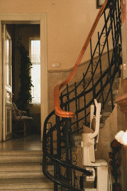 Inside a residential property, a staircase with a curved wooden handrail and ornate black wrought iron balustrade leads upward. At the base of the staircase, a white stair lift is attached to the steps, positioned for use in home relocation or moving processes. The surroundings include a nearby open doorway revealing a room with a window covered by curtains, allowing natural light to illuminate the space. A wooden chair with a upholstered seat is visible beside the doorway, and a potted plant can be partially seen further inside. The interior has beige walls and marble flooring, with a radiator beneath the window. The overall setting reflects a typical indoor environment during furniture transport or packing and moving activities, with no additional objects or wrapped furniture visible in the scene.