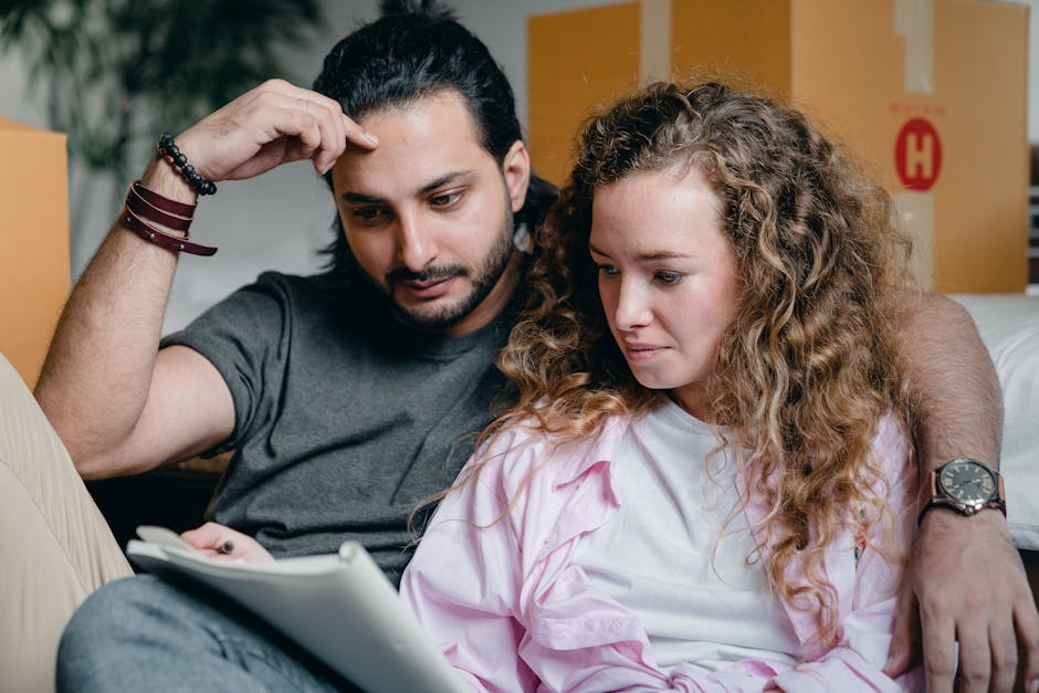 Inside a property with several packed cardboard boxes stacked in the background, a man with dark hair and a beard, wearing a grey T-shirt, is sitting with his arm around a woman with curly blonde hair