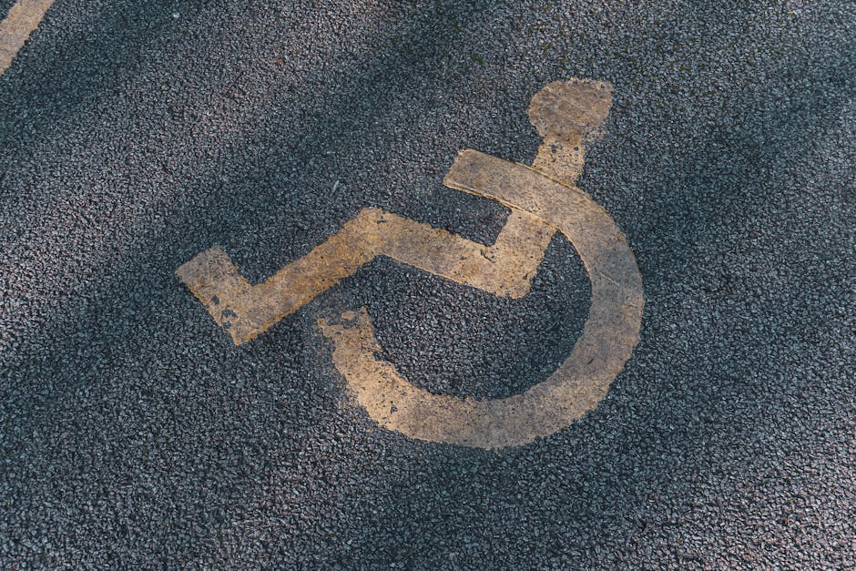 A close-up view of a weathered yellow parking space marking on dark asphalt, featuring a painted wheelchair symbol indicating accessible parking. The symbol shows a stylised figure seated in a wheelchair, with the figure's head, arms, and legs visibly outlined. The surface of the asphalt has a rough texture with small pebbles, and the paint appears worn and slightly faded, suggesting frequent use over time. This marking is located outdoors, possibly near an entrance or parking lot adjacent to a property, providing guidance for mobility-impaired access. It is visible in an area prepared for a home relocation or furniture transport, with the surrounding environment implying proximity to a residential or commercial building. The presence of this accessible parking space underscores consideration of parking logistics during house removals and furniture transport, as managed by Merton Movers.
